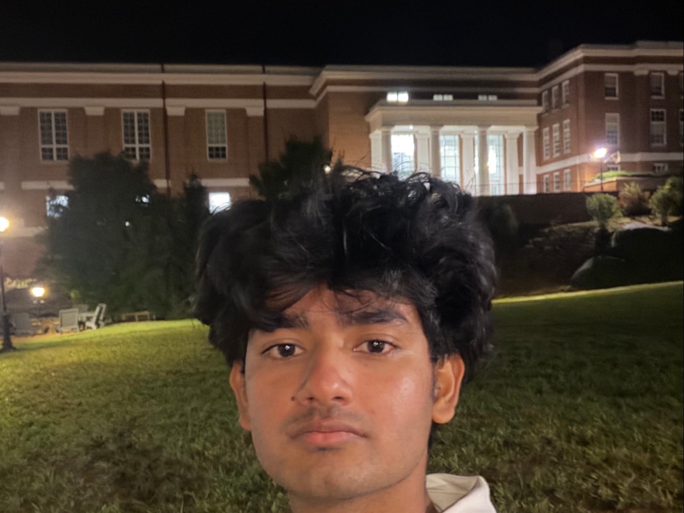Aditya taking a selfie at night on a lawn, in front of a lit brick building with white columns.